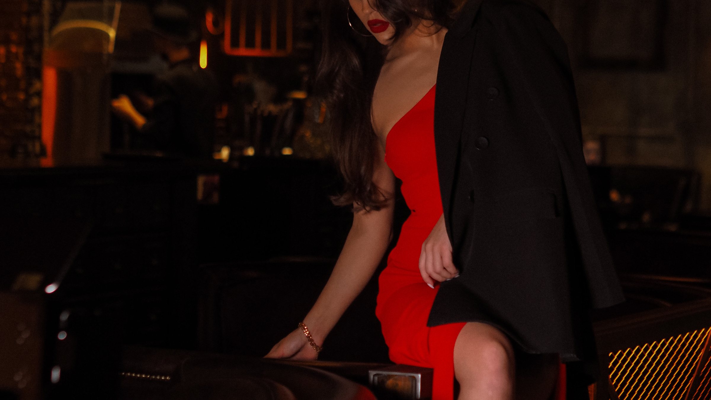 A woman in a red dress and black blazer perched at a dim bar